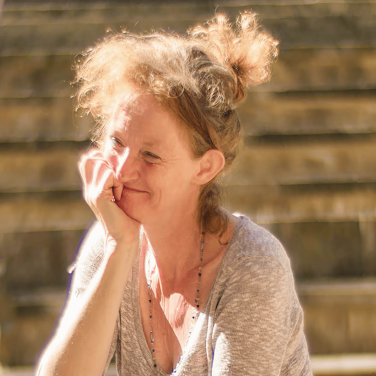 Kelly Hunter MBE smiling warmly in sunlight, seated outdoors with her hand to her face. Founder of Flute Theatre, she develops groundbreaking Shakespeare adaptations for autistic individuals and neurodivergent audiences.