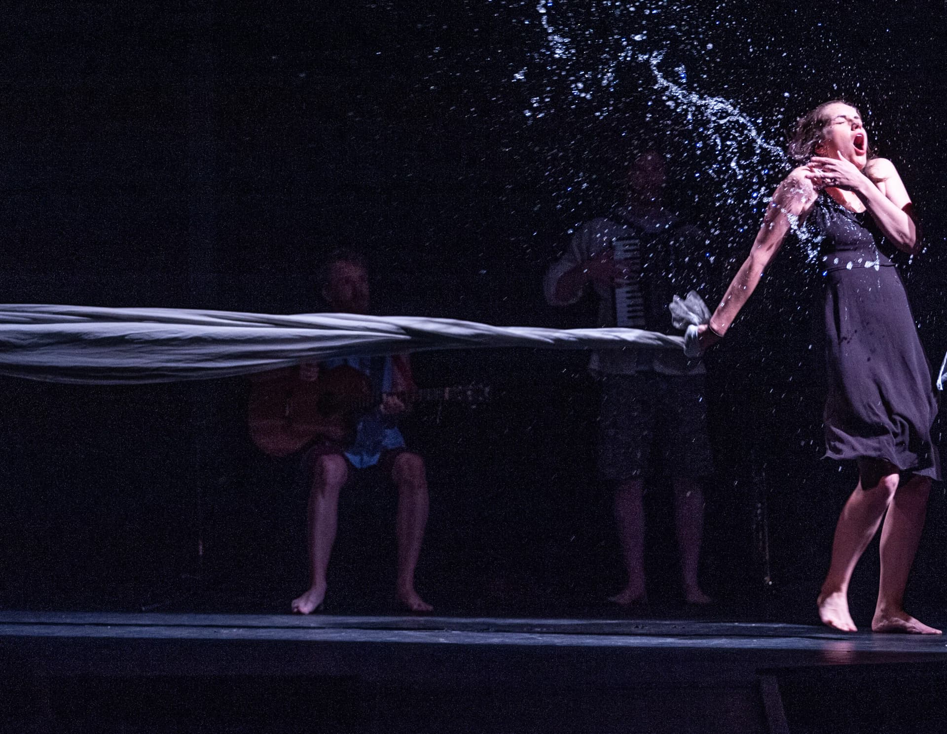 A performer in a black dress is dramatically splashed with water on stage, reacting with surprise and intensity, while musicians play guitar and accordion in the dimly lit background.