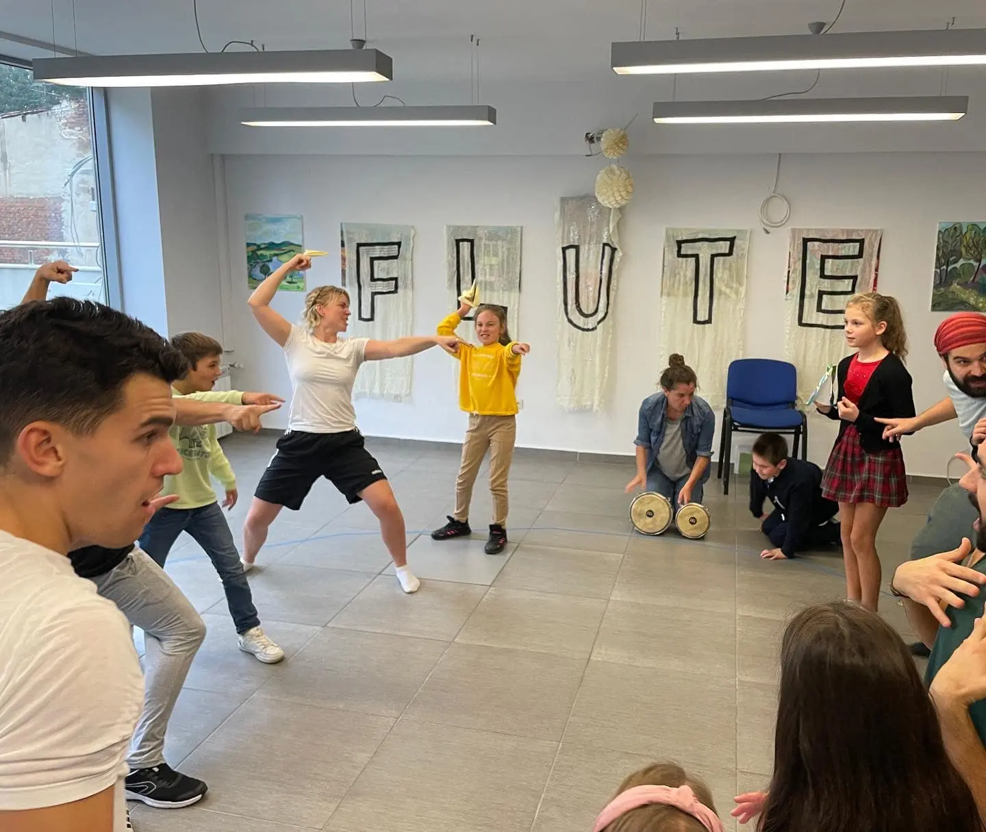 Participants engage in a lively, interactive retelling of Pericles. A facilitator in white energetically mirrors a young girl in a yellow sweatshirt as others join in, pointing dramatically. Behind them, fabric banners spelling FLUTE hang on the wall, setting the immersive scene for this Pericles workshop.