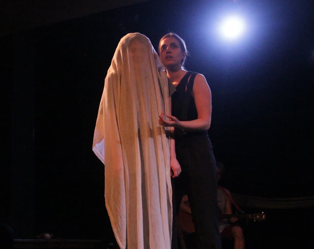 A woman in a black outfit stands beside a figure draped completely in a white sheet like a ghost, lit by a bright stage light behind them.