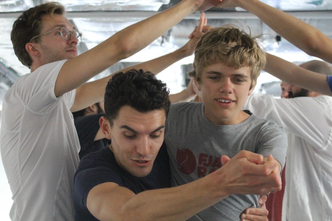 A young boy and an adult man engage in a dance activity with raised arms while other participants move in the background of a light-filled studio.