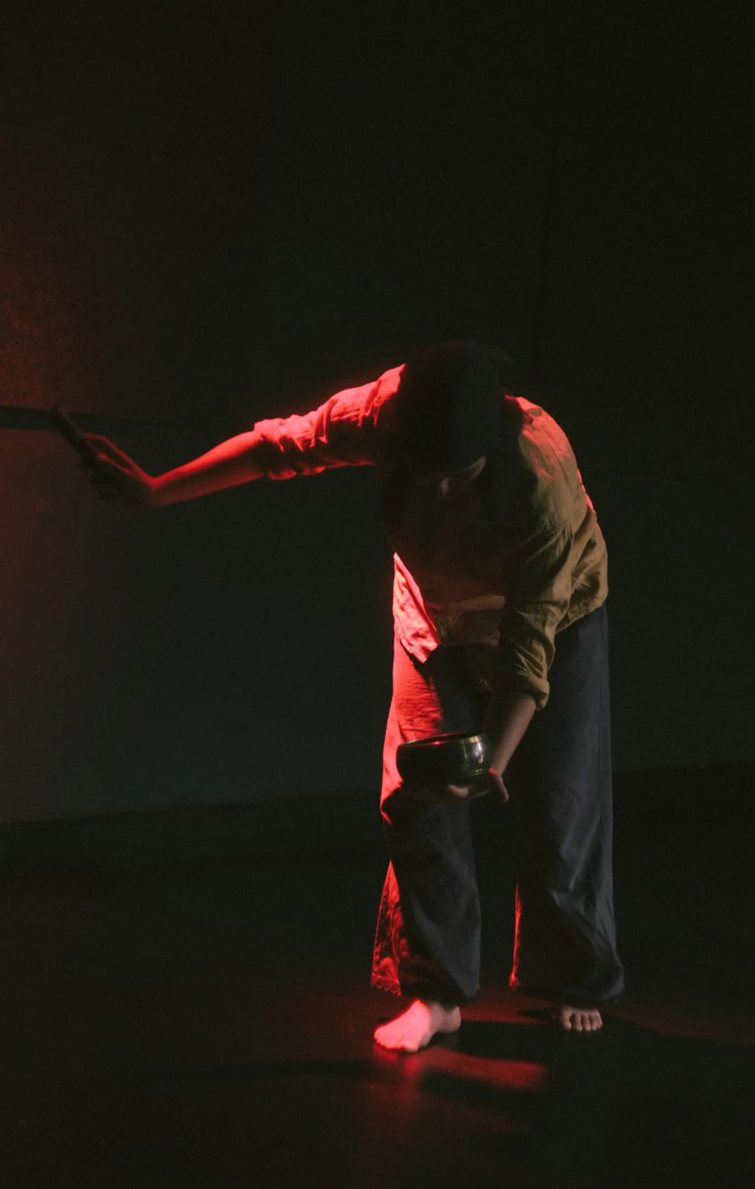 A performer in loose clothing is illuminated by red stage light while striking a metal bowl, creating a ritualistic and intense atmosphere.