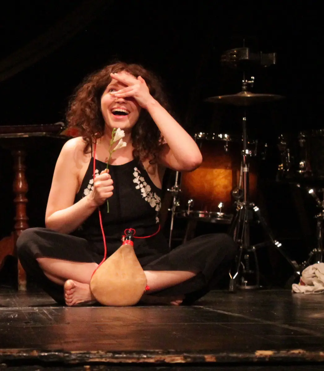 A woman with curly hair sits cross-legged on stage, holding a flower and a gourd flask, smiling brightly with one hand playfully covering part of her face.