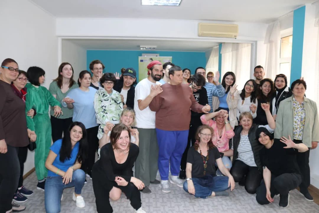 A large group of participants, including autistic individuals, families, and performers, smiling and posing together in a bright indoor space.