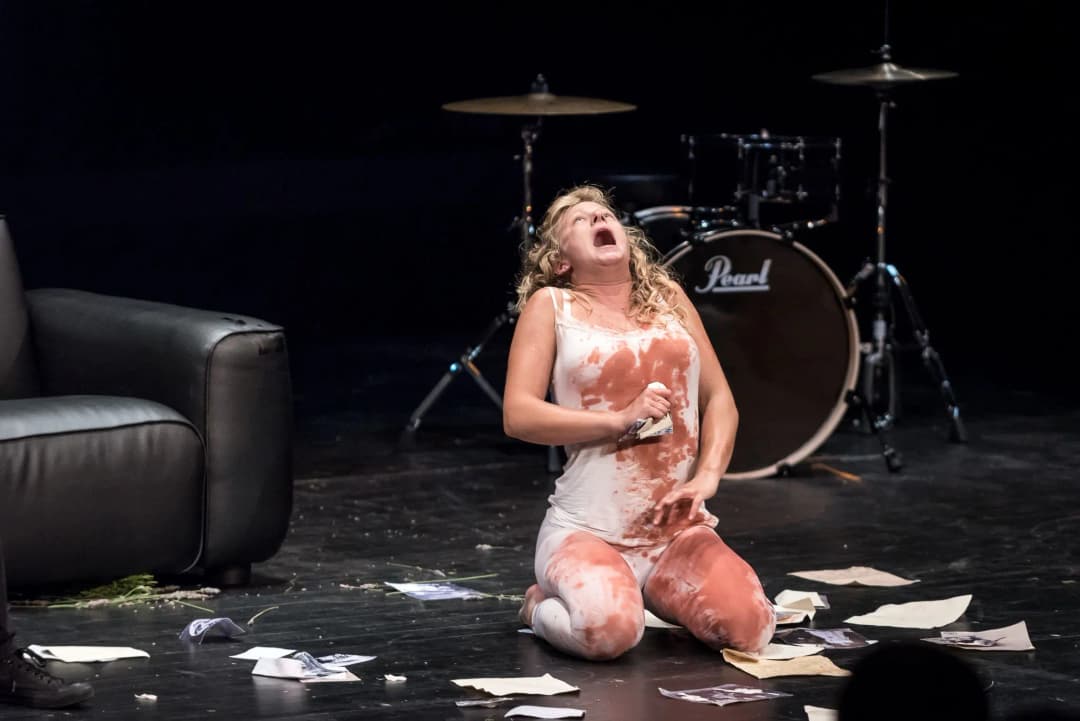 An actor in a white outfit kneels on stage, covered in fake blood, screaming in anguish amidst scattered papers, a drum set visible in the background.