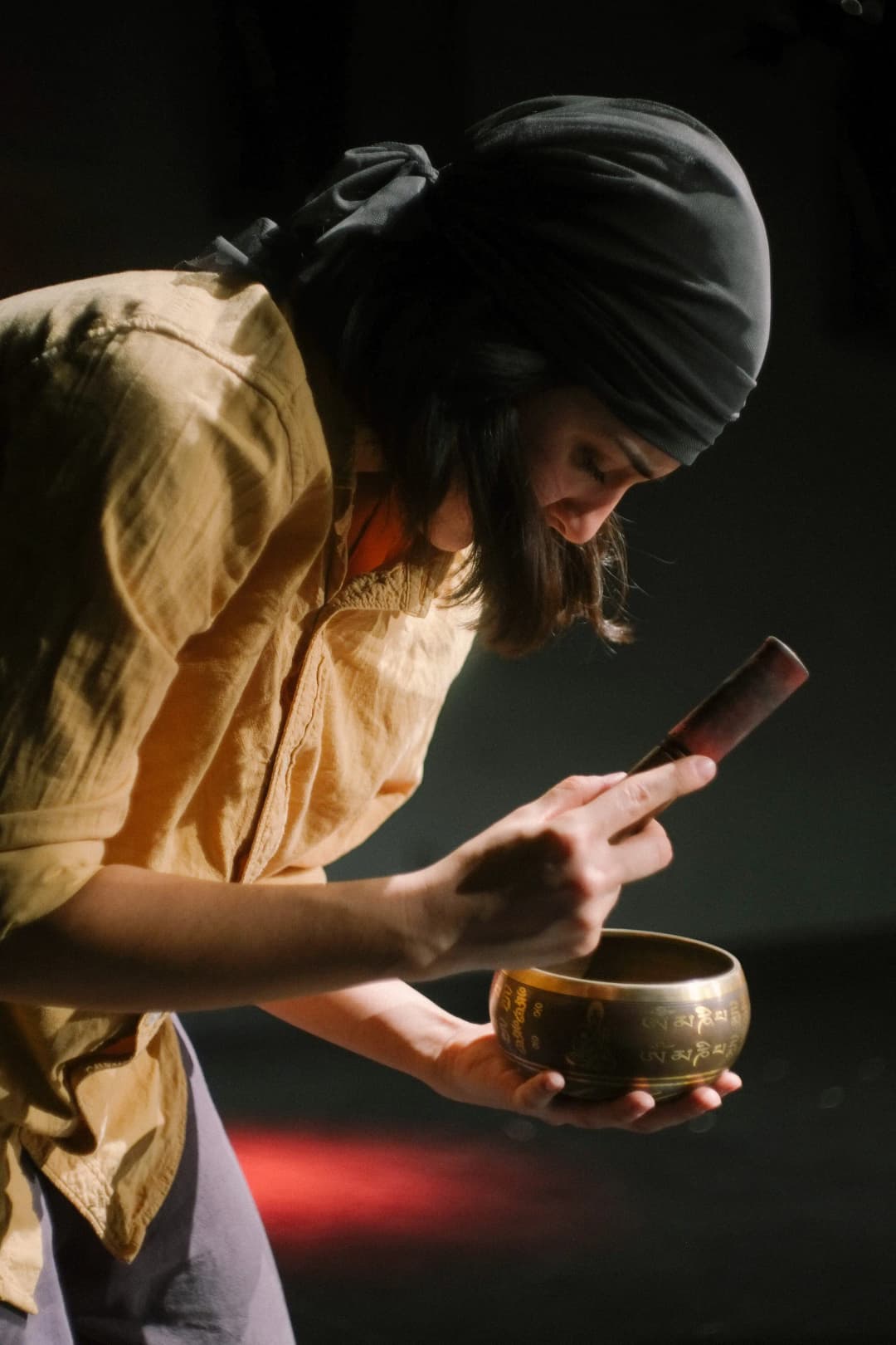 A close-up of a performer focused on playing a singing bowl, bathed in soft stage lighting that highlights their concentration.