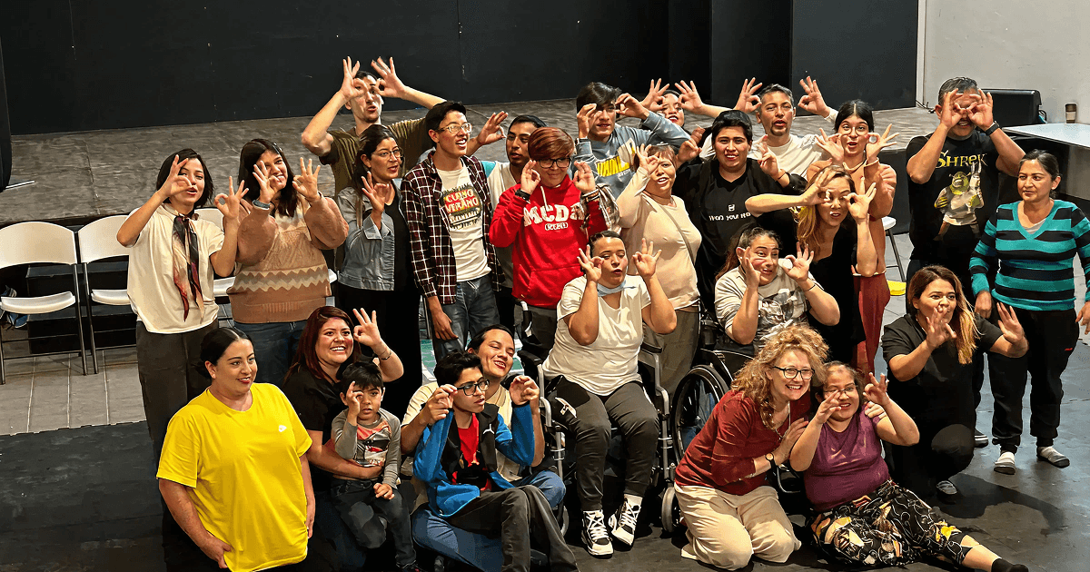A joyful group photo of Flute Theatre actors, disabled participants, and local facilitators at the Otros Territorios Festival in Mexico. Everyone is smiling and making playful hand gestures, celebrating the end of an inclusive theatre workshop.