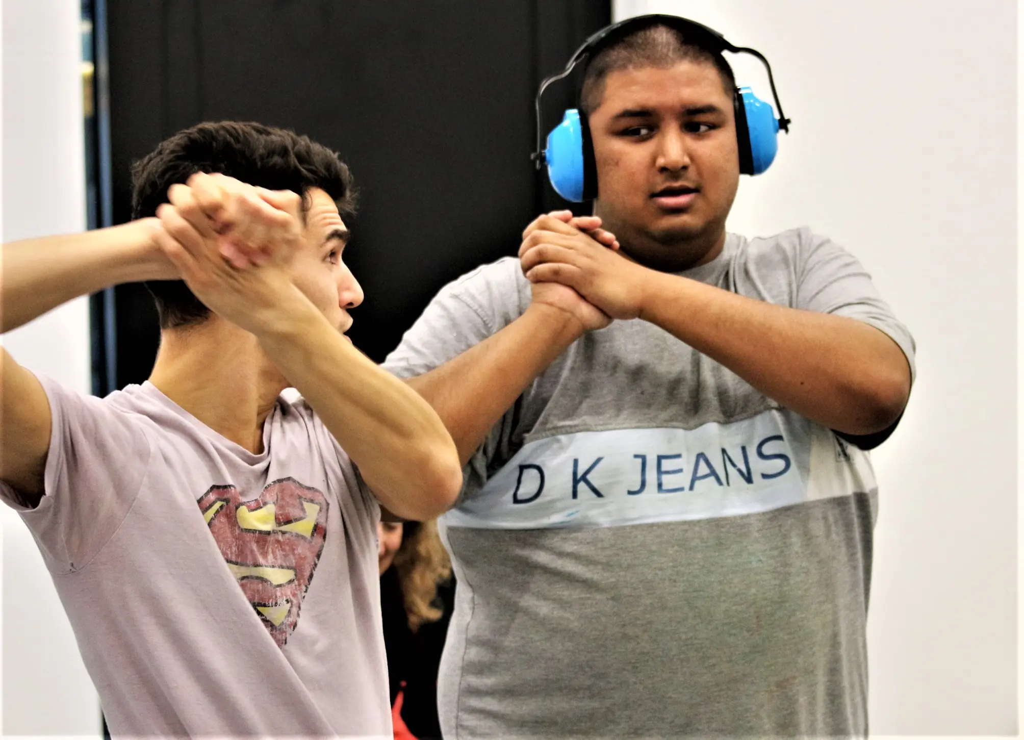 Two participants clasp hands while dancing in a Brighton inclusive theatre workshop, one wearing blue ear defenders and a grey DK Jeans shirt, the other in a pale Superman T-shirt.