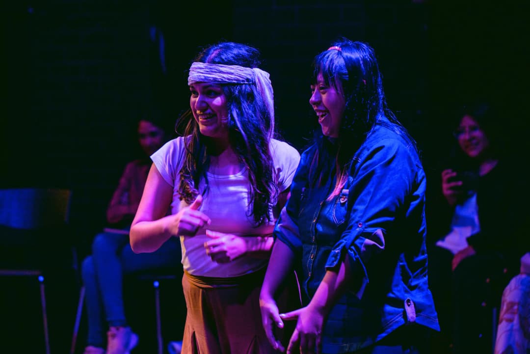 Two actors smiling and reacting with joy on stage during an interactive theatre piece in Peru, with the audience visible in the background.