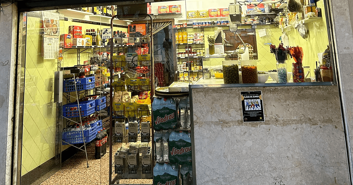 Small corner shop in Montbau, Barcelona with shelves stacked full of bottled drinks, snacks, and preserved goods; a window display features jars of olives and sweets, evoking everyday life in the neighborhood where Flute Theatre works with profoundly autistic individuals.