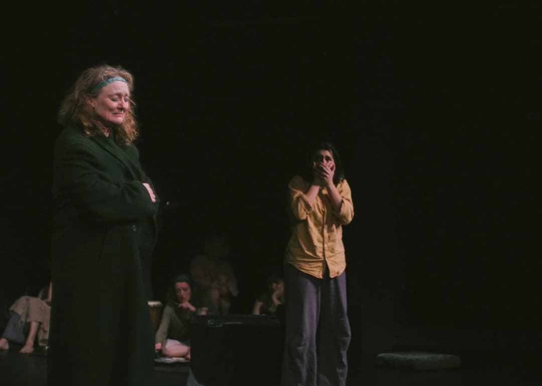 A woman wearing a dark coat and a headband looks emotional, while another performer in a yellow shirt reacts with shock, covering her mouth with her hands.