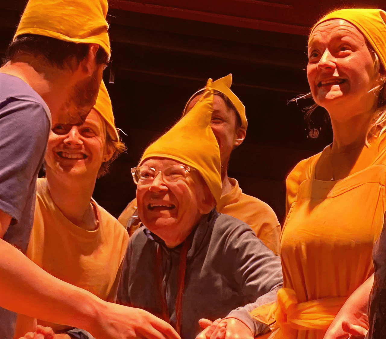 A heartwarming moment from a Flute Theatre production shows a group of actors and an older autistic participant wearing matching yellow hats and costumes. They are smiling and holding hands in a shared moment of joy. This sensory-friendly Shakespeare adaptation uses the Hunter Heartbeat Method to foster emotional connection and inclusion, welcoming autistic individuals of all ages into the performance experience.