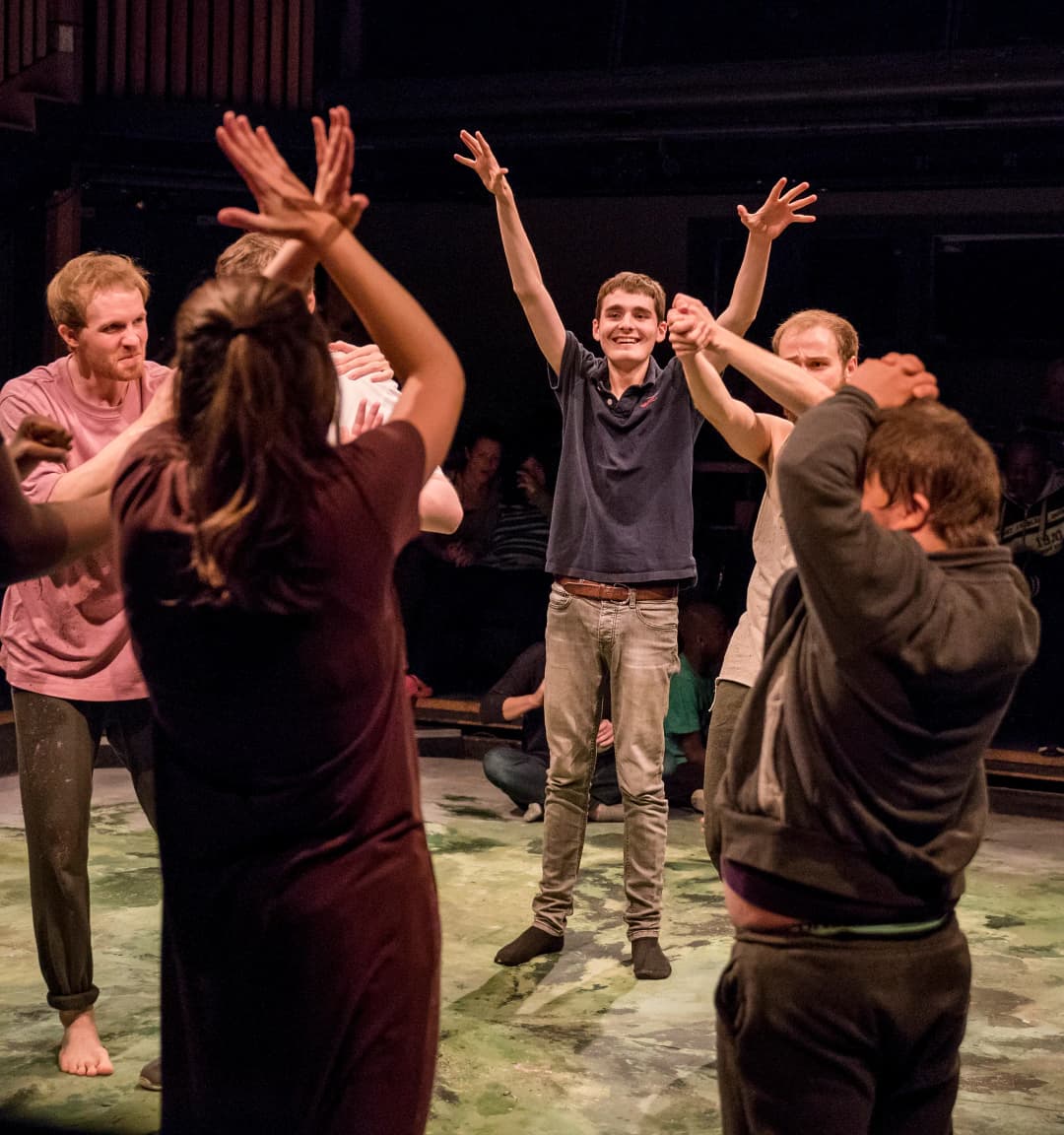 A group of participants, including neurodiverse individuals, stand in a circle on stage with joyful expressions. A young man in the center raises his arms high in excitement, while others around him join in the playful movement.