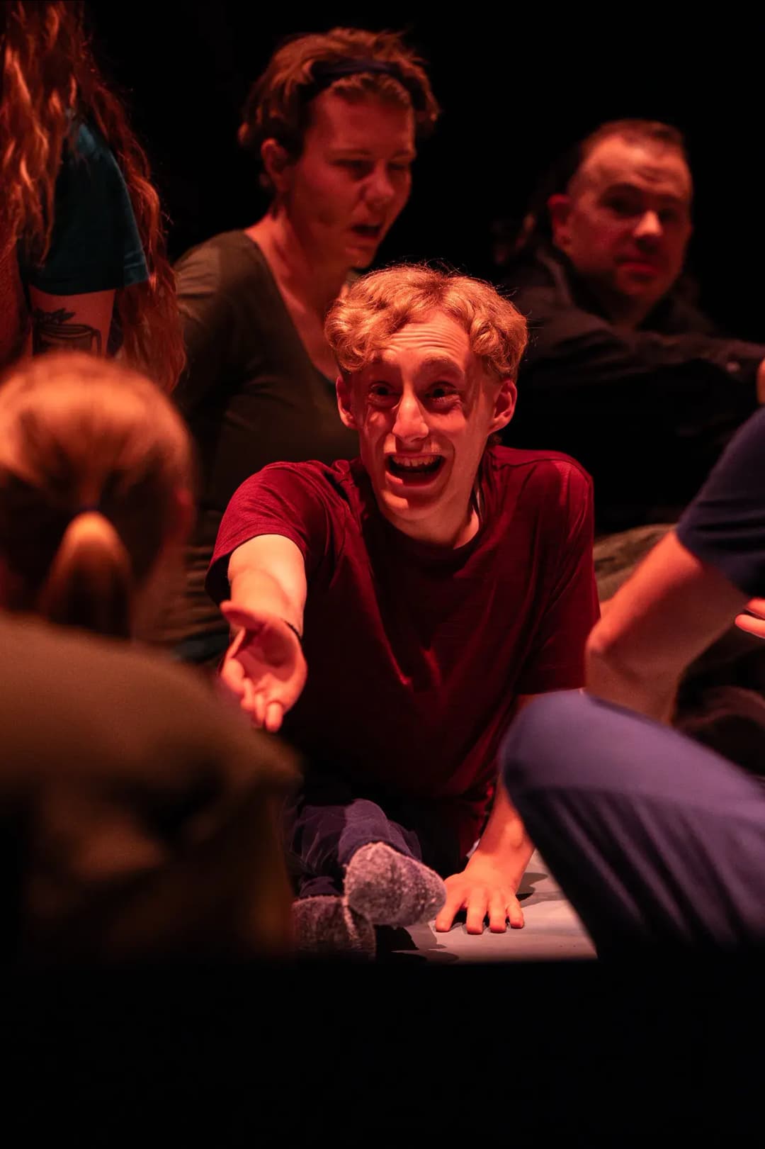 A young man with blonde hair smiles brightly, reaching out his hand toward another participant while others look on warmly.