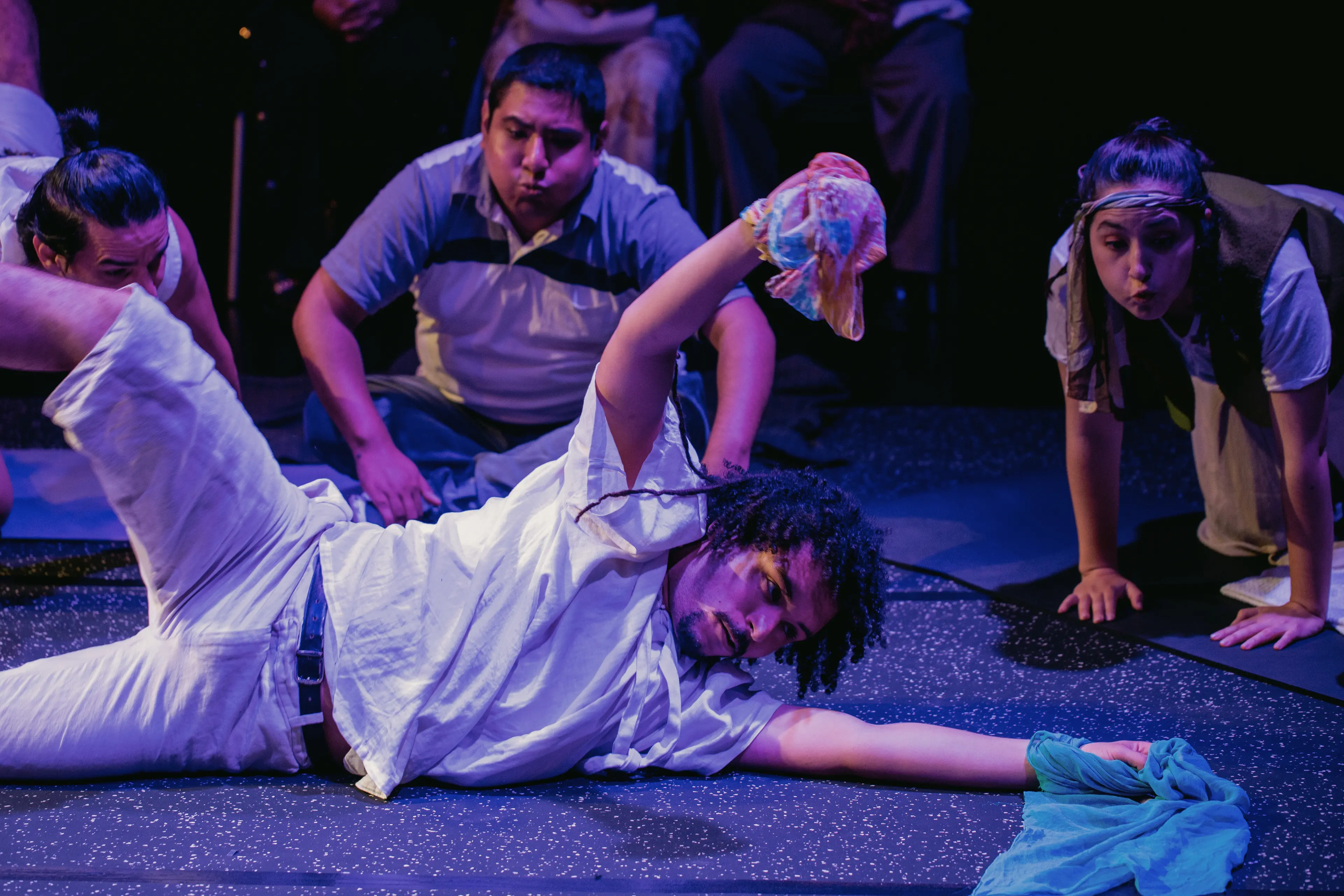 A performer lies dramatically on the floor, holding colorful fabric, while other cast members crouch and watch intently, enhancing the sensory-rich performance.