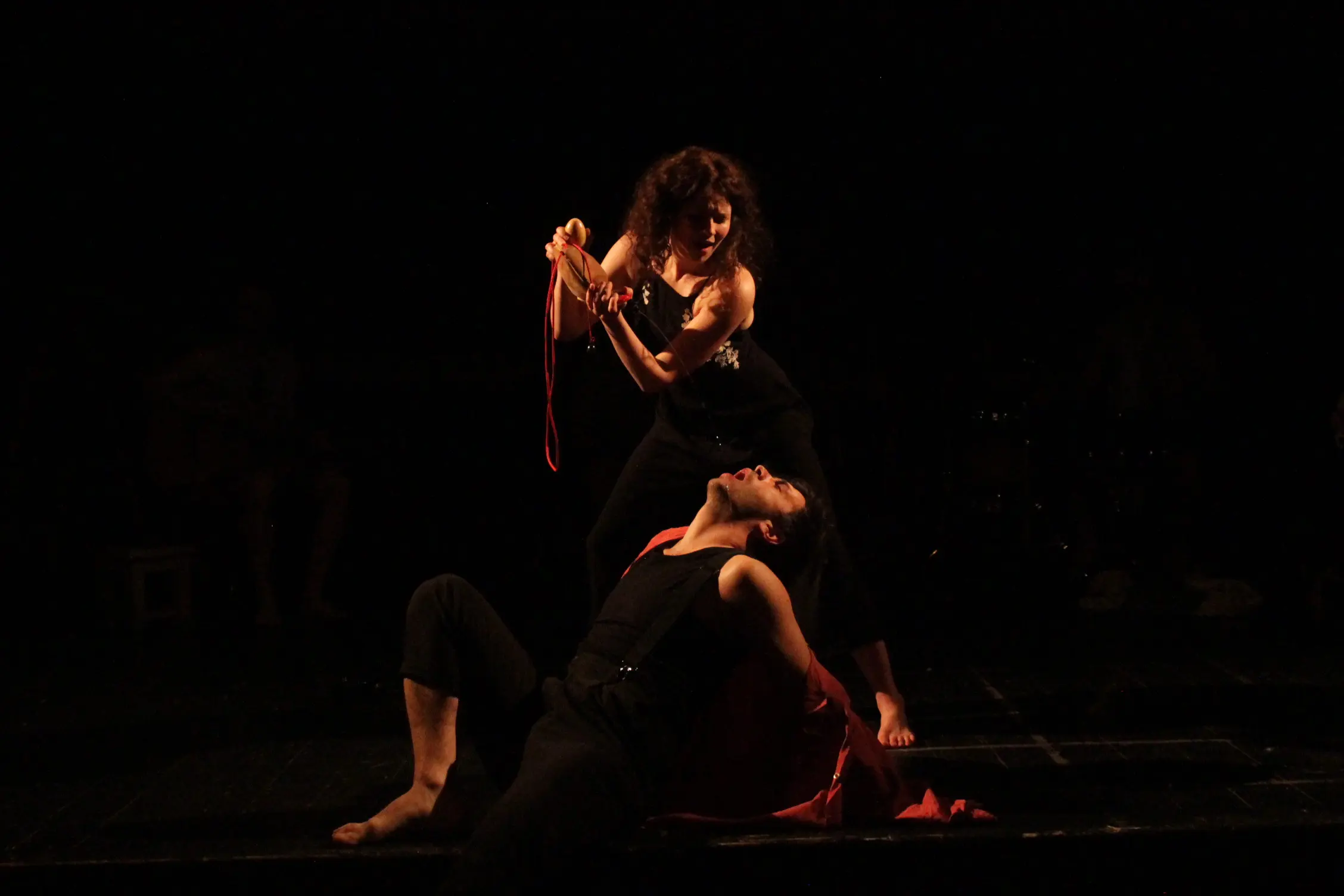 A performer holds a red cord and gourd above another actor dressed in black, who leans back dramatically with a red cloth draped around them, evoking a sense of tension and struggle.