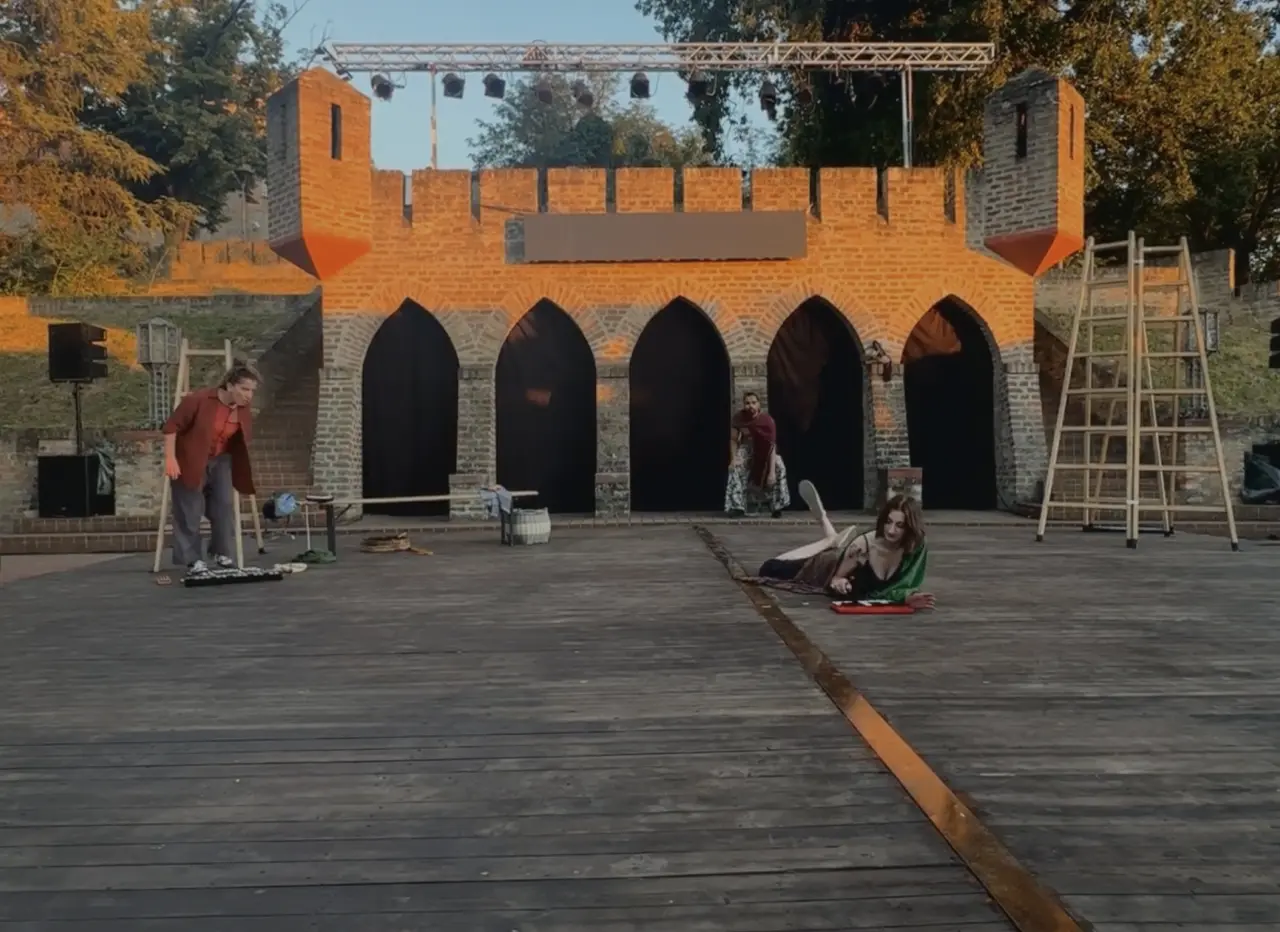 Actors performing A Midsummer Night’s Dream on an outdoor stage for the Shakespeare Festival Novi Sad Serbia 2024 with a medieval castle-like backdrop, one lying on the wooden floor in the foreground, another adjusting props, and a third in the distance near the arched set entrances.