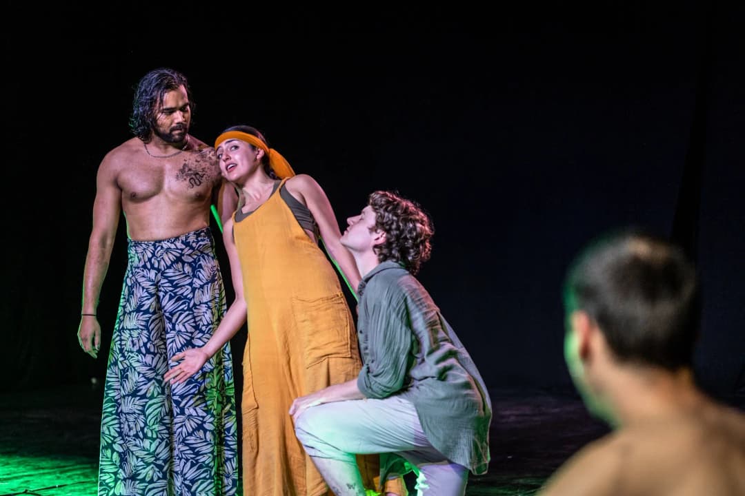 Three actors on stage performing A Midsummer Night’s Dream, with one in patterned trousers standing tall, another in a yellow dress leaning towards a kneeling actor, all set against a dark backdrop with dramatic green lighting.