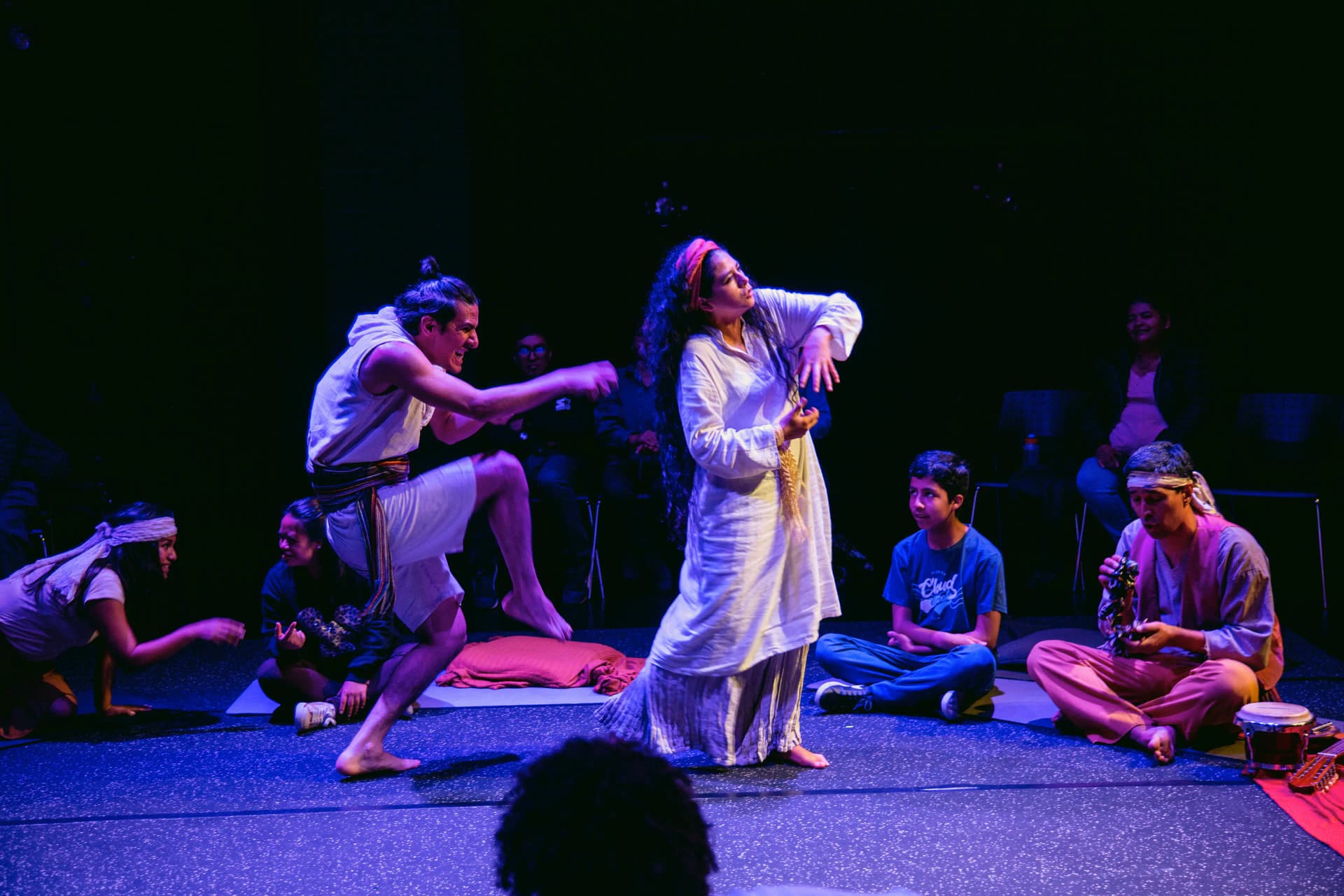 Actors in dynamic motion performing a theatrical scene, surrounded by seated audience members, in an intimate theatre setting in Peru.