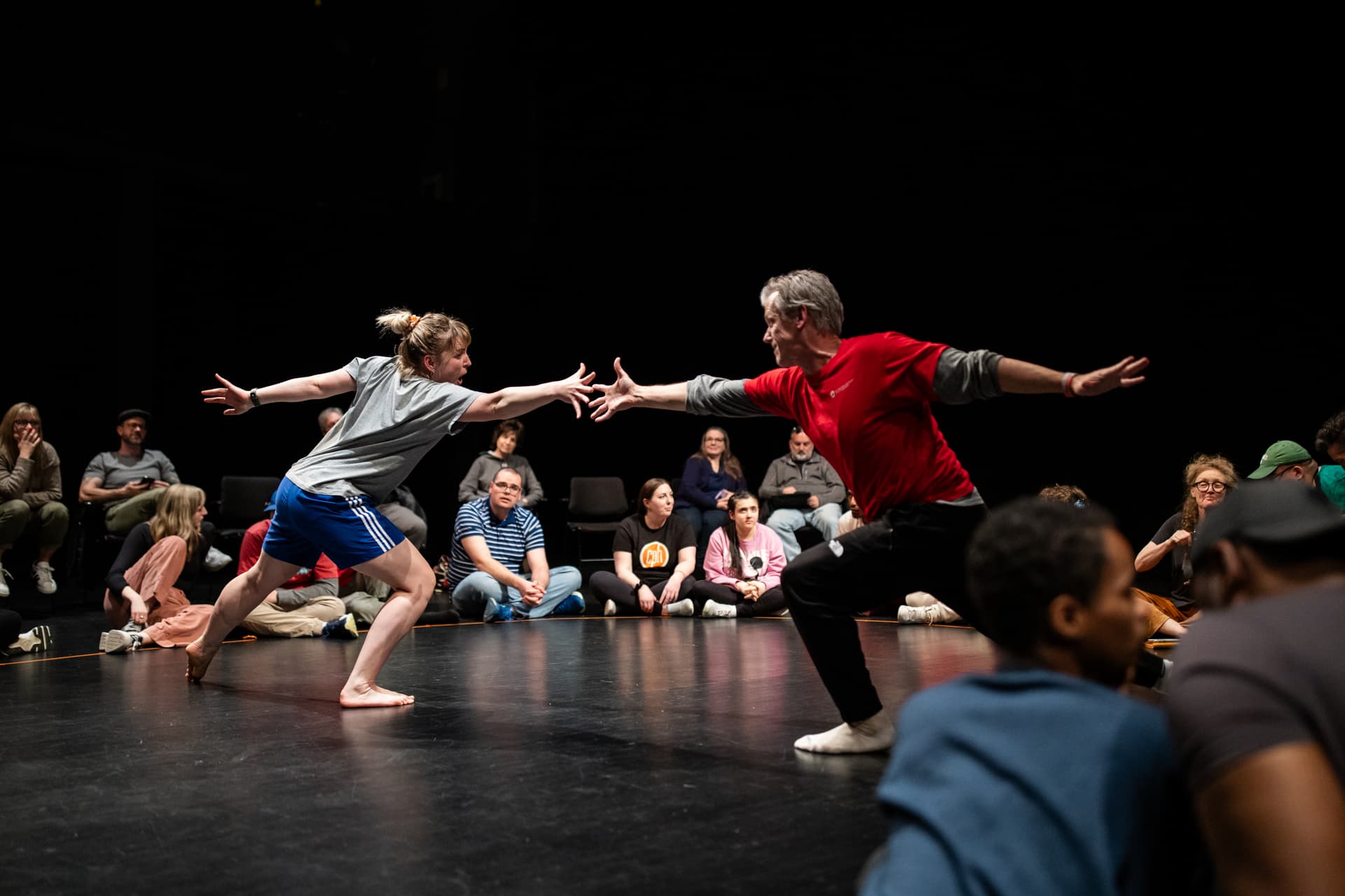 Two performers, a woman in blue shorts and grey t-shirt and a man in a red t-shirt and black trousers, reach toward each other mid-movement in an expressive, dynamic pose. They are surrounded by an audience seated on the floor in a circle, closely watching the interactive performance in an intimate, inclusive theatre setting.