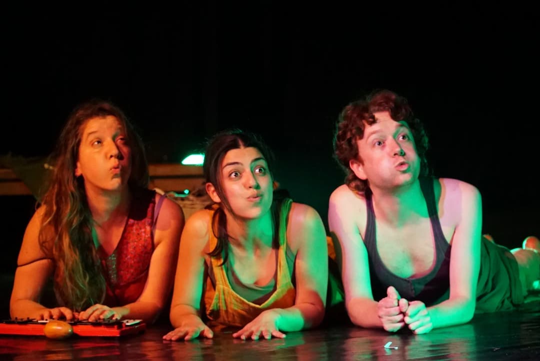 Three actors lying on the stage floor during A Midsummer Night’s Dream, with playful, exaggerated expressions as they engage with the audience, illuminated by green stage lights.