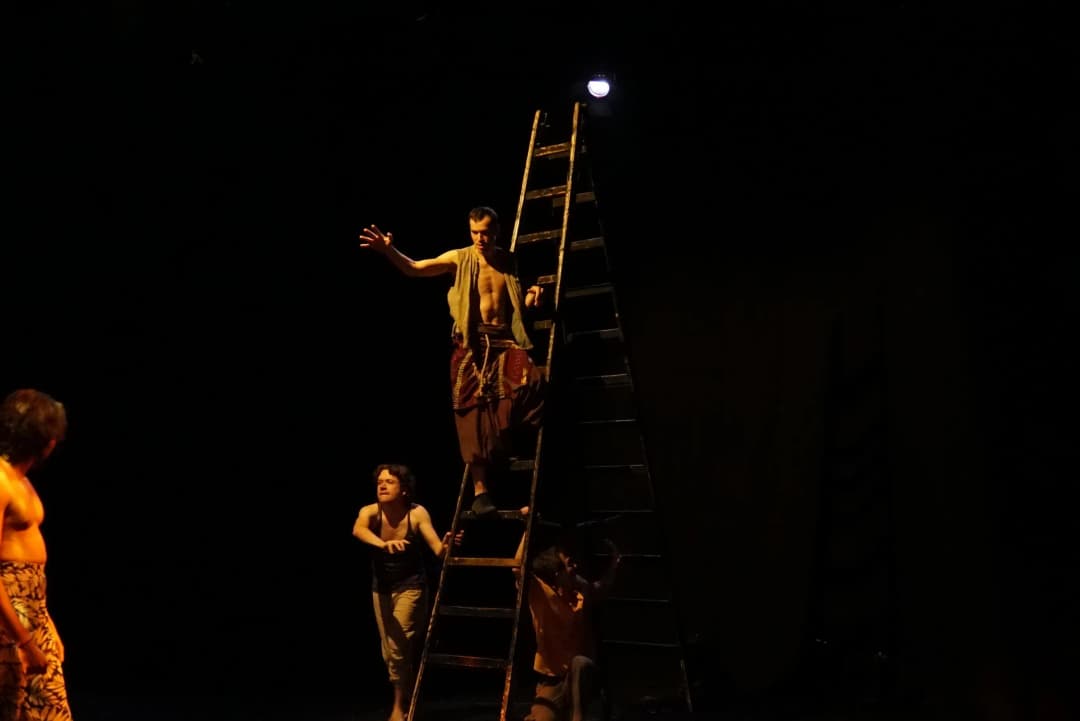 An actor balanced on a tall ladder reaching out dramatically during A Midsummer Night’s Dream, while others look on and interact below, all in dim, moody lighting.