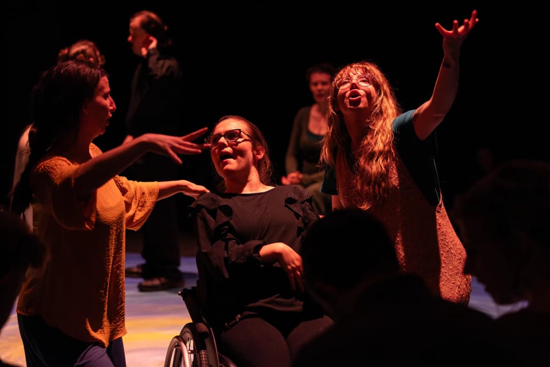 Three performers, including one in a wheelchair, sing and gesture expressively, illuminated by warm stage lighting as others watch.
