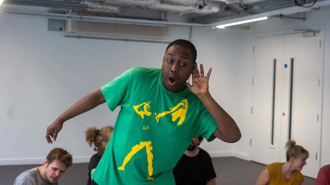 A man in a bright green t-shirt strikes a dramatic pose with one hand cupped to his ear, as if listening closely, while others sit and watch in the background of a studio.