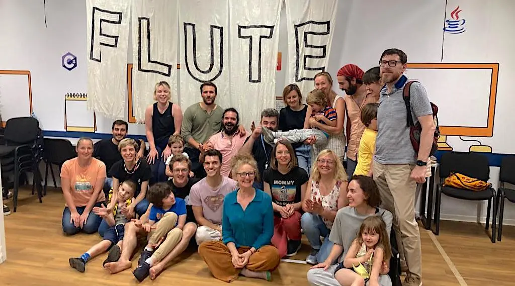 A group of adults and children smiling together in front of a large banner with the word "FLUTE" written on it, in a bright indoor space with chairs and colorful wall art.