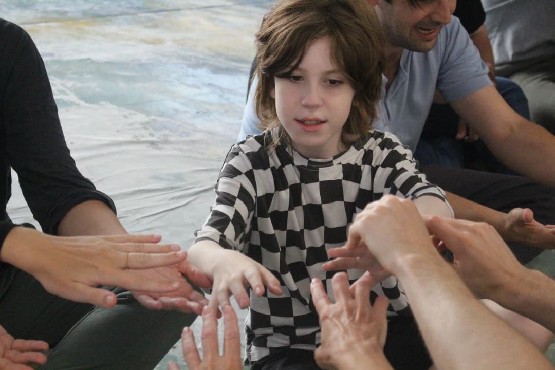 A child in a black-and-white checkered shirt focuses intently while gently touching hands with multiple people in a group exercise.