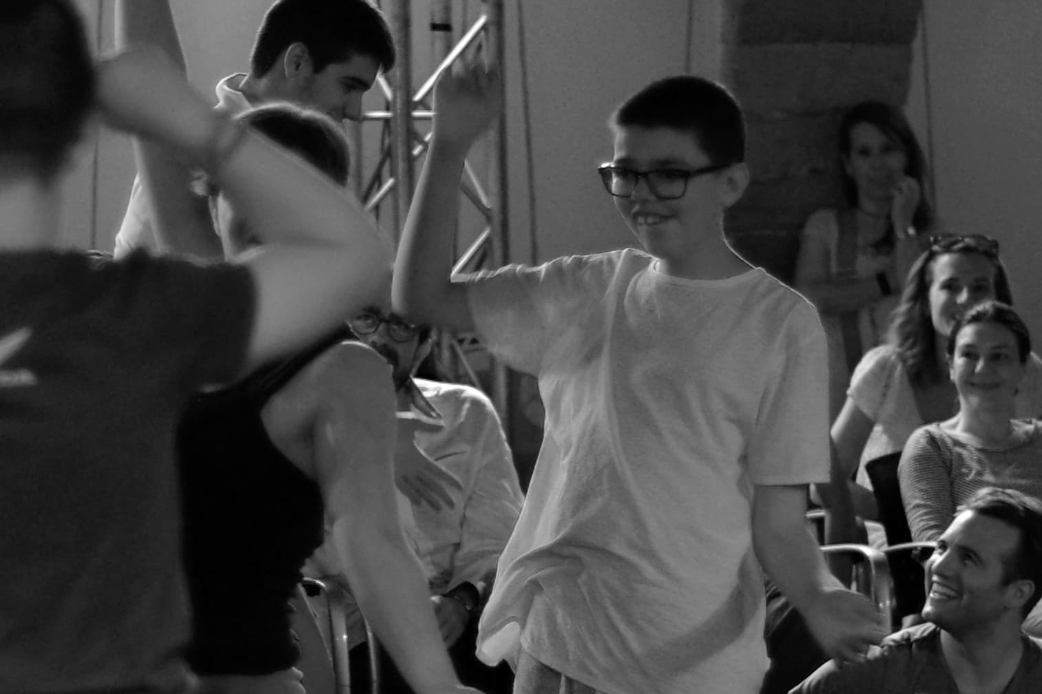 A young boy wearing glasses smiles and raises his arms in movement during an inclusive performance or workshop in Granollers, 2019, while others around him watch with joy and encouragement.