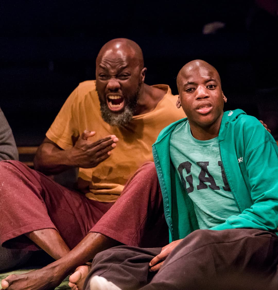 Two men sitting on the floor, one with a green hoodie and another older man in a yellow shirt, both animatedly expressing emotions as part of the interactive performance.