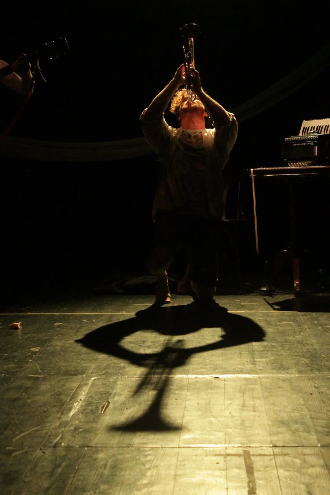 A musician kneels under a spotlight, lifting a trumpet high above their head, casting a large, dramatic shadow on the stage floor.