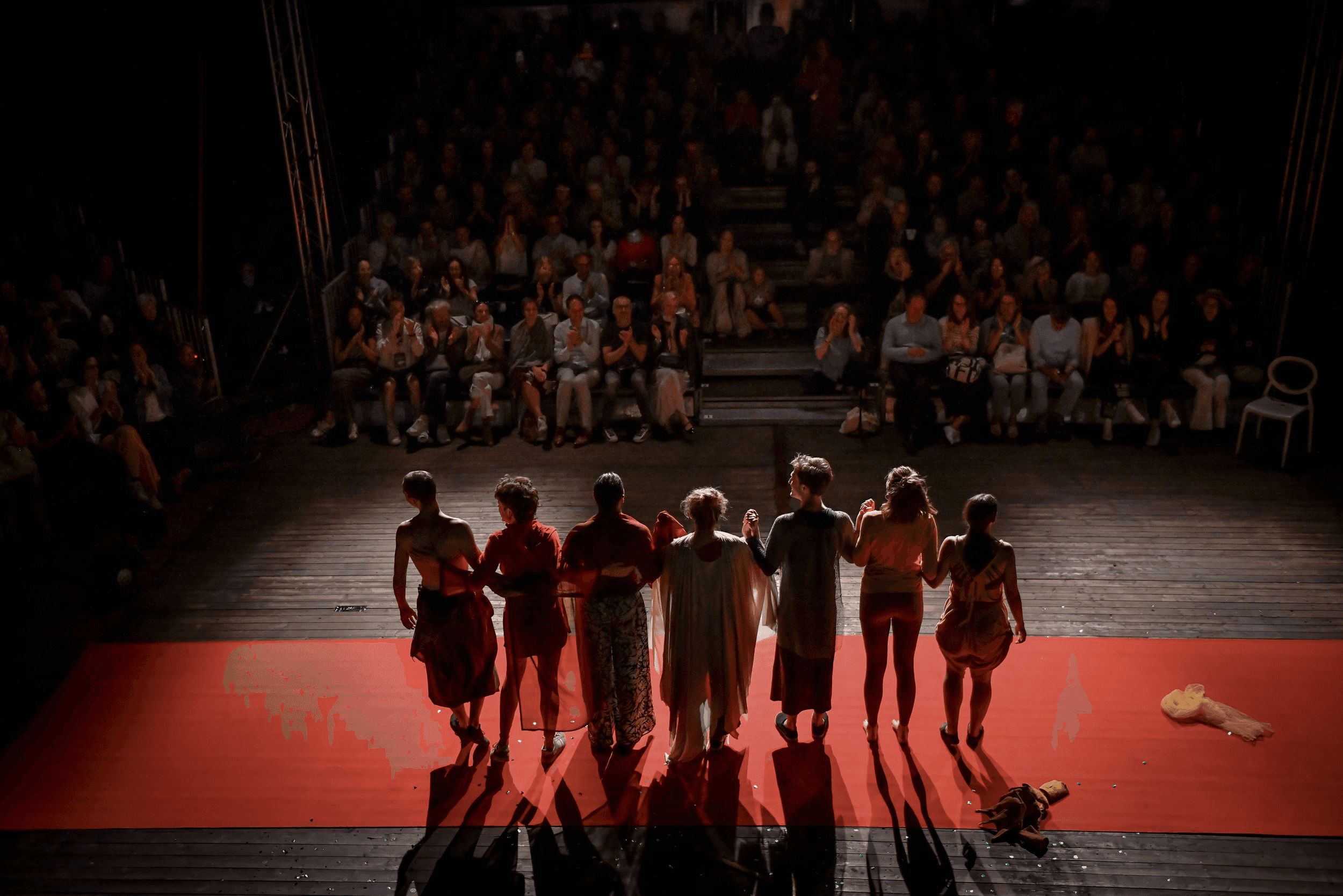 The ensemble of Flute Theatre stands arm-in-arm on a red carpet, bowing to a full house after an inclusive adaptation of Shakespeare. The diverse cast, lit warmly from behind, is silhouetted against an applauding audience. This image captures the culmination of a sensory-rich performance designed for autistic individuals and their families, using the Hunter Heartbeat Method to create meaningful, interactive theatre experiences.