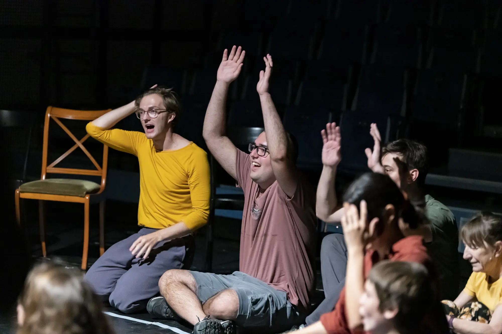 Audience members and performers sit on the floor with raised hands, smiling and reacting together in an interactive moment.