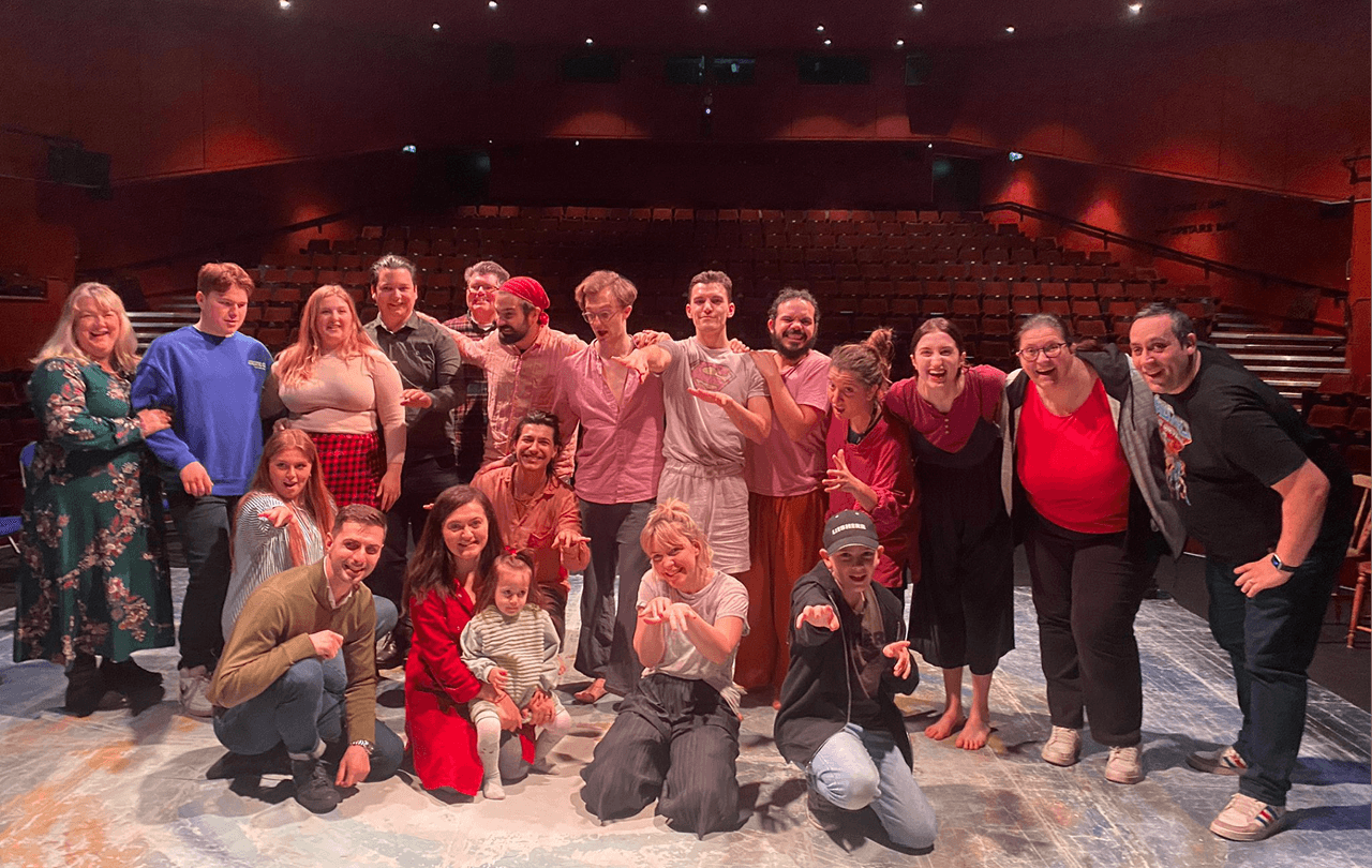 A large inclusive group of actors, participants, and facilitators from Flute Theatre gather on stage after a performance. The group, including autistic individuals and neurodivergent families, smiles and poses together in a celebratory moment following an interactive Shakespeare adaptation.