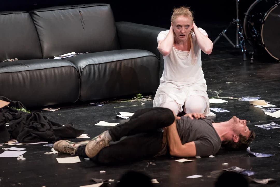 A distraught actress in white kneels with her hands over her ears beside a man lying on the stage, clutching his chest, surrounded by scattered papers and debris.