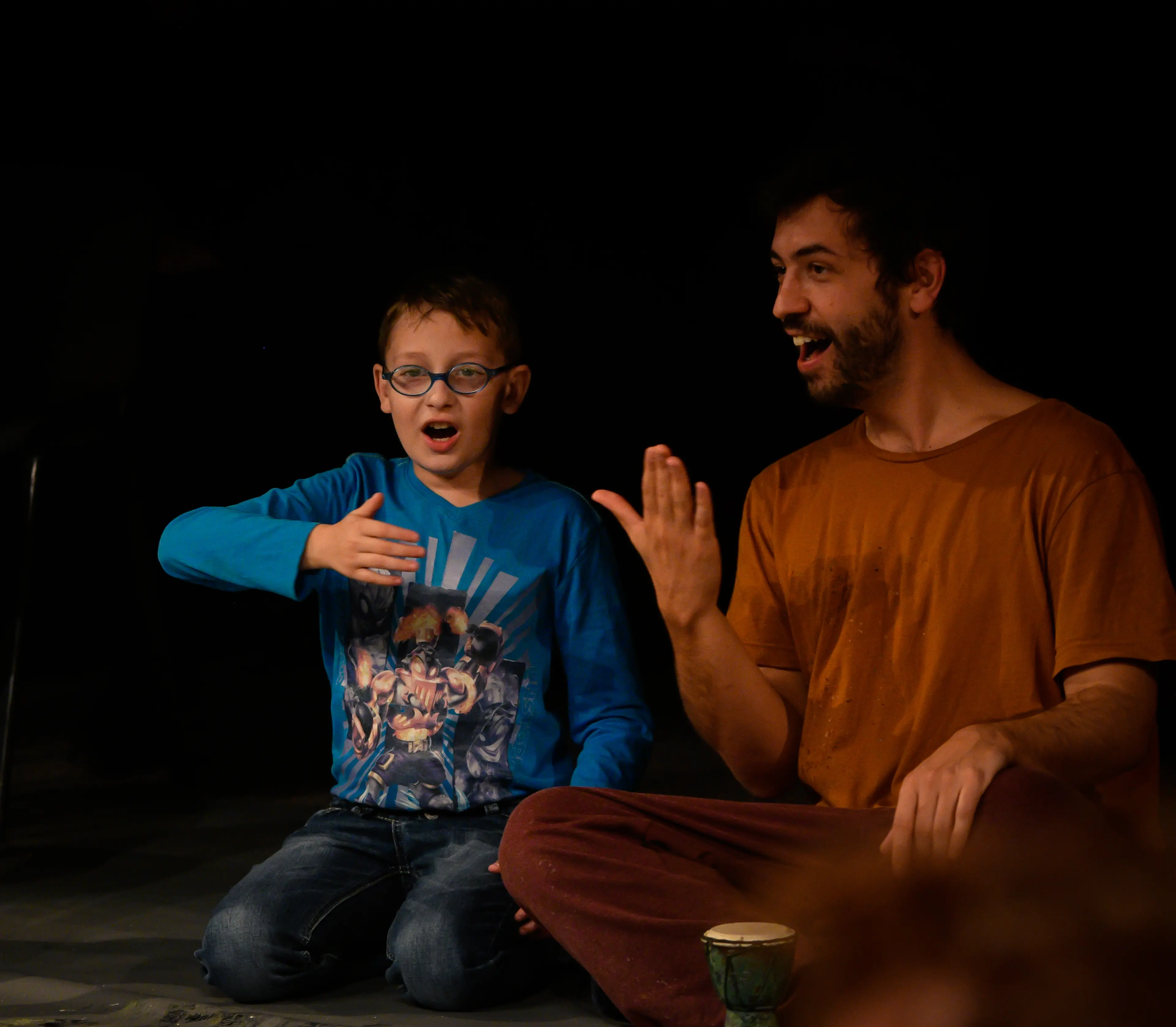A young boy in glasses and a performer in an orange shirt sit cross-legged on the floor, enthusiastically gesturing with their hands as they engage in the performance.