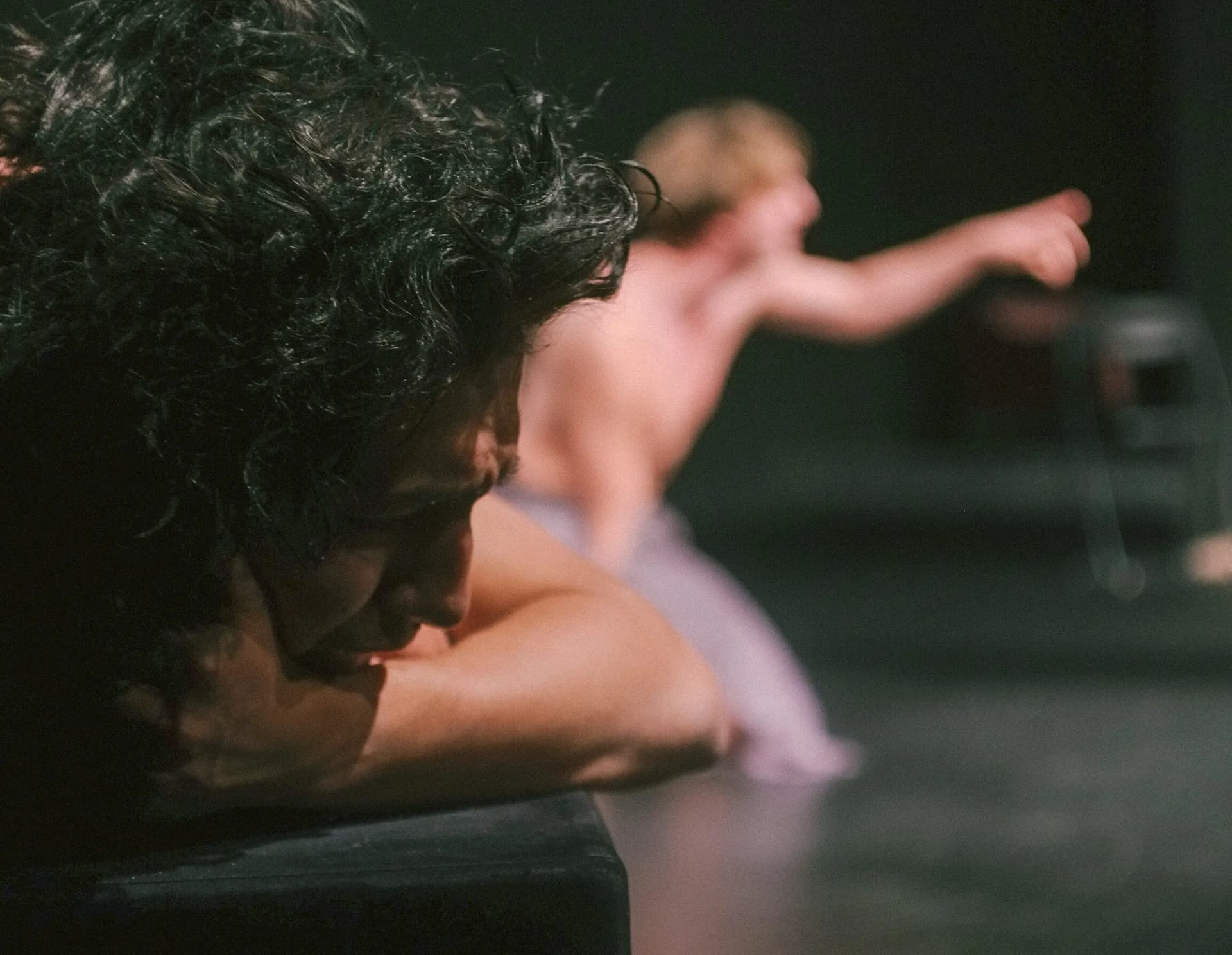 A shirtless actor sits on the ground with a surprised expression as another shirtless performer looms behind, hands raised in a dramatic, menacing gesture.