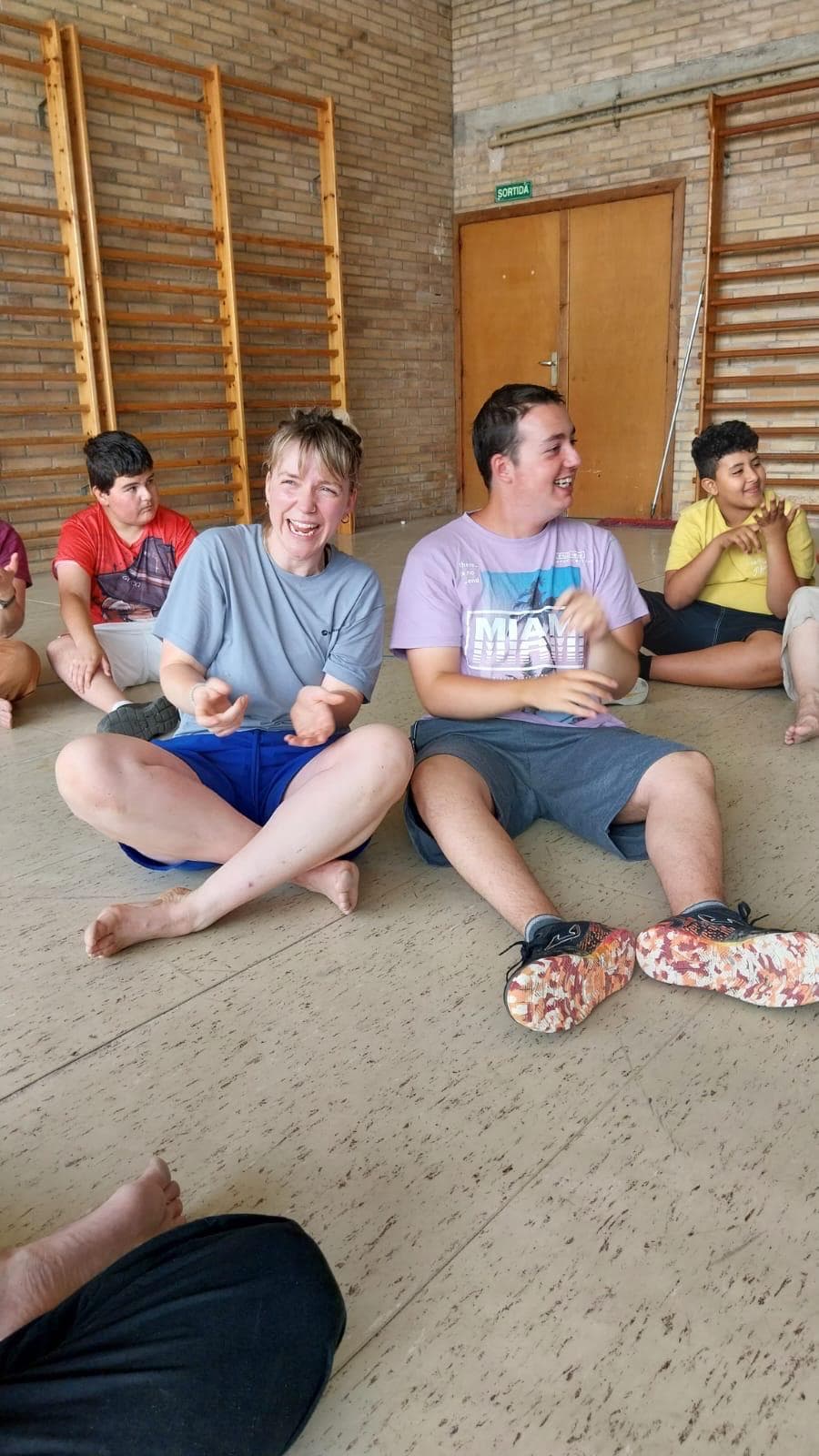 Actor and autistic participant laughing and clapping during a Flute Theatre session at Centre d’Educació Especial Montserrat Montero in Granollers, Catalunya. They sit barefoot on the gym floor alongside friends, sharing a moment of connection through Shakespeare-inspired play.