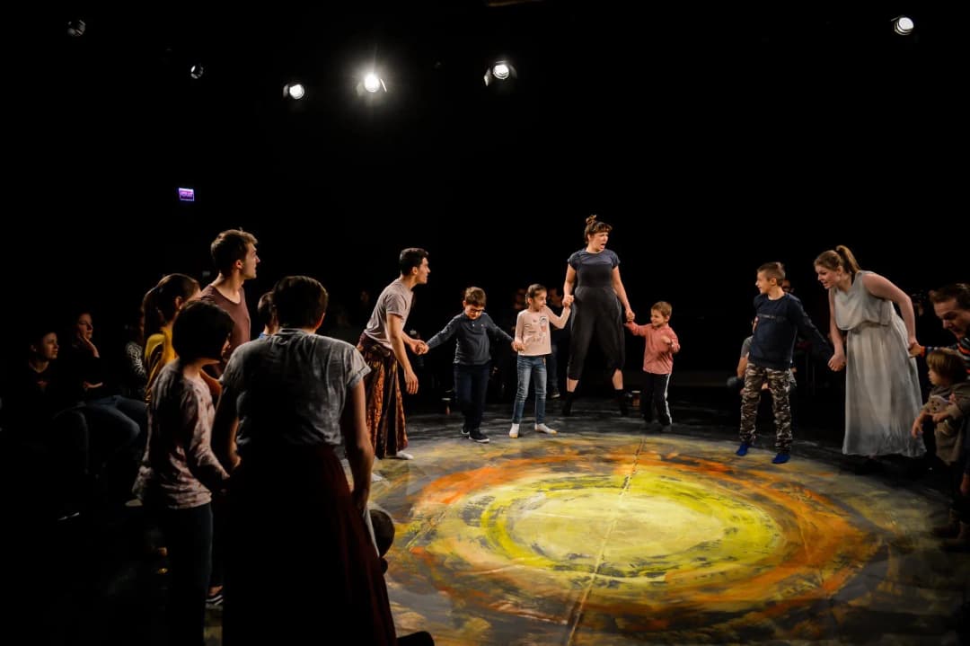 A group of children and adults holding hands in a circle during a theatre workshop, jumping together on a colorful stage floor.