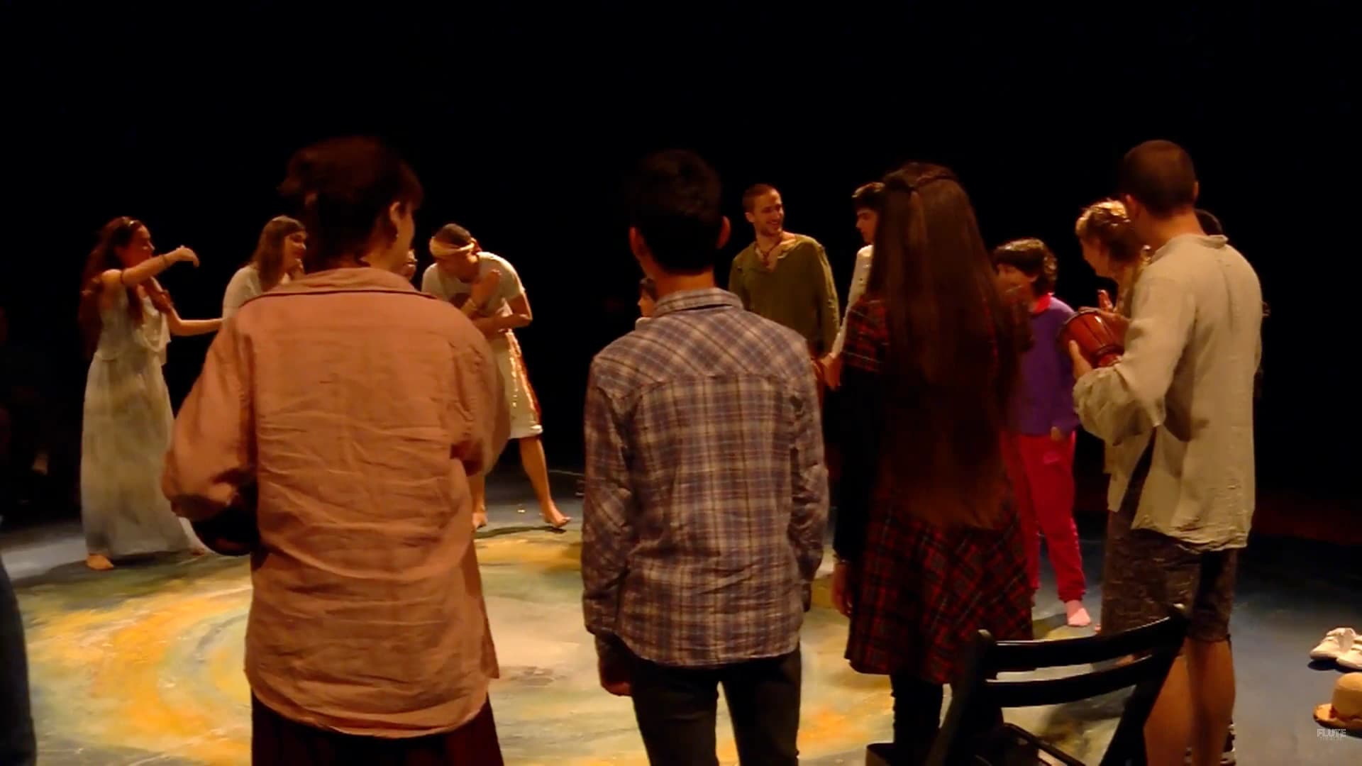 A group of performers and participants stand in a circle on a dimly lit stage, engaging in an interactive moment of La Tempesta in Barcelona (2017), surrounded by soft, warm lighting and a painted floor design.