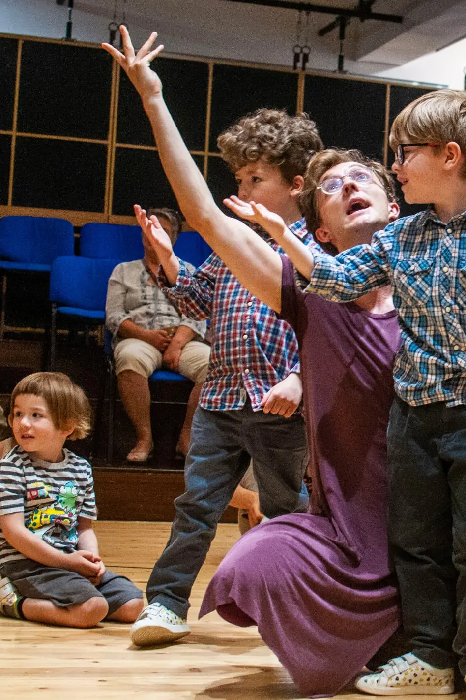 Josh, the performer, kneeling dramatically with arms raised, engaging two young boys in plaid shirts while another child watches nearby in a cozy indoor performance space.
