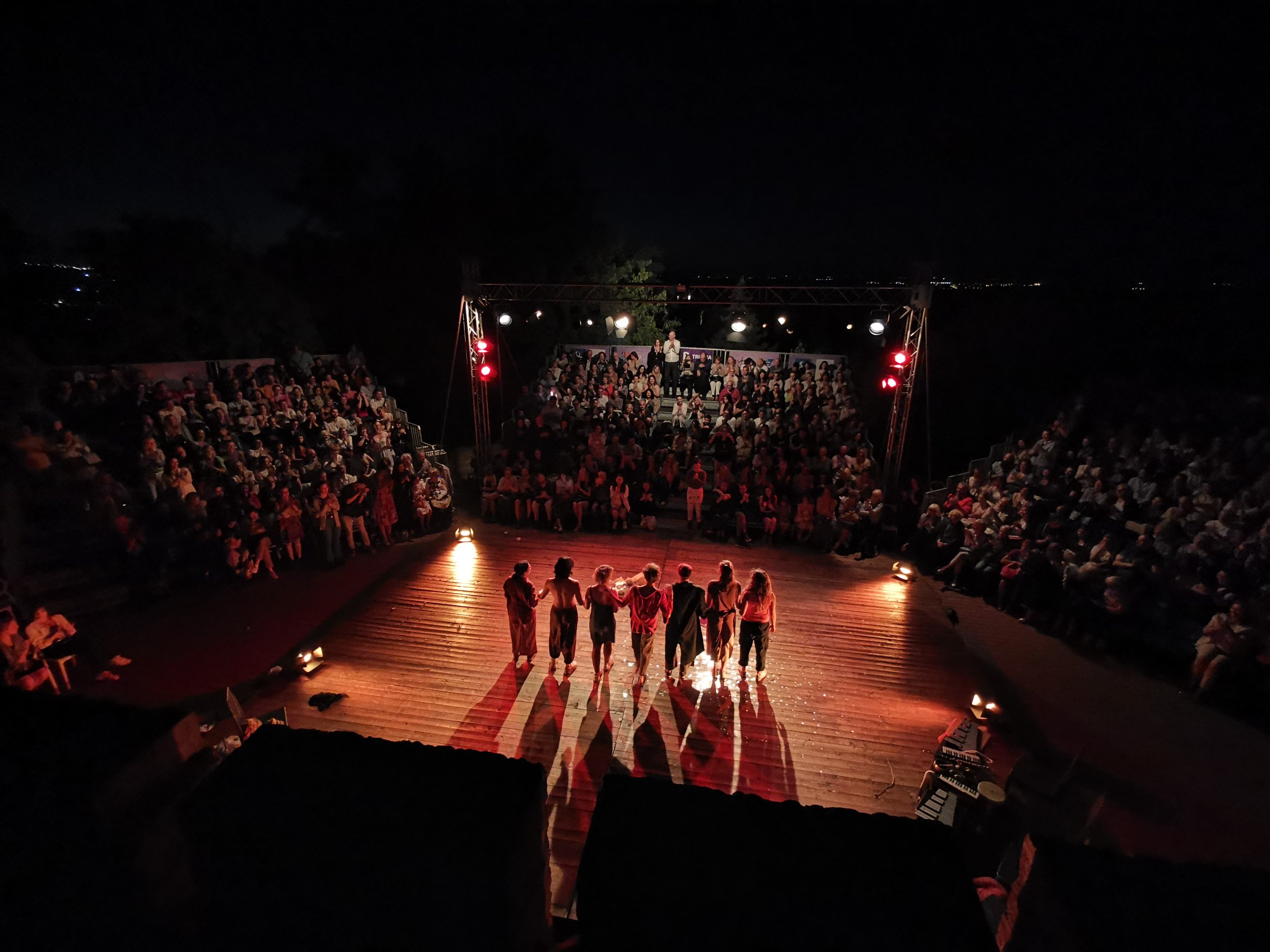 A wide-angle nighttime photograph of a Flute Theatre performance in an outdoor amphitheatre, with a group of actors standing center stage in a close circle, illuminated by warm stage lighting. The large audience surrounds them in a semi-circle, fully engaged with the performance. Though this particular event was not an adaptation for autistic individuals, it reflects Flute Theatre’s broader mission to create inclusive, emotionally resonant Shakespearean experiences. Flute Theatre also leads workshops and training programmes in the Hunter Heartbeat Method for actors, educators, applied theatre students, and families around the world.