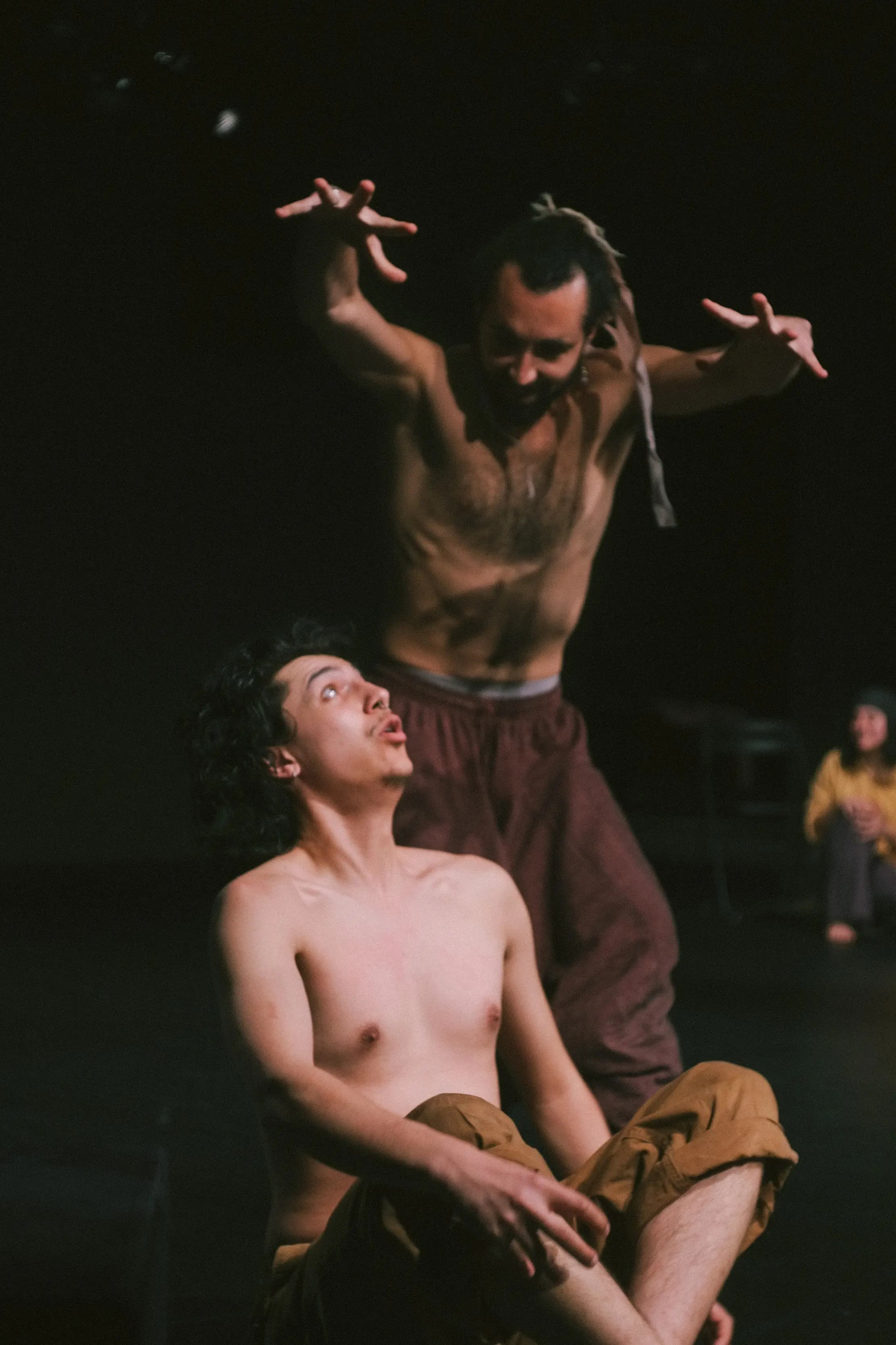A close-up of a performer leaning on their arm, looking distressed, while another blurred figure in the background points accusingly on stage.