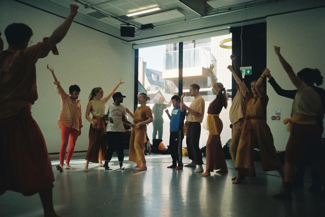 Participants in colourful costumes raise their arms in unison during an inclusive theatre rehearsal in Brighton, with natural light streaming through large studio windows.