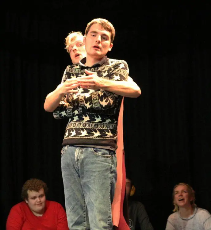A performer stands at the front of the stage in Brighton, with another person behind them wrapping their arms gently around their chest in an expressive movement, while others watch in the background.