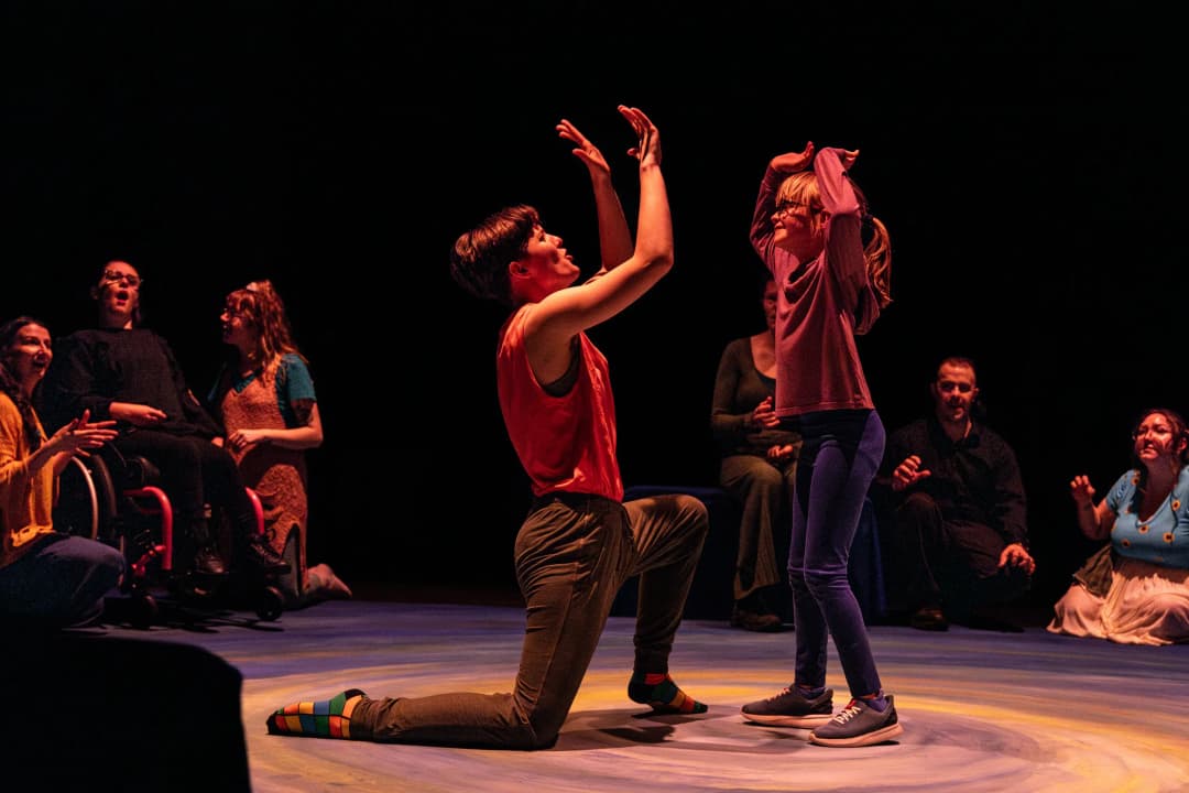 An adult performer kneels joyfully, raising their arms while a young girl mimics the gesture, surrounded by smiling participants watching the interaction.