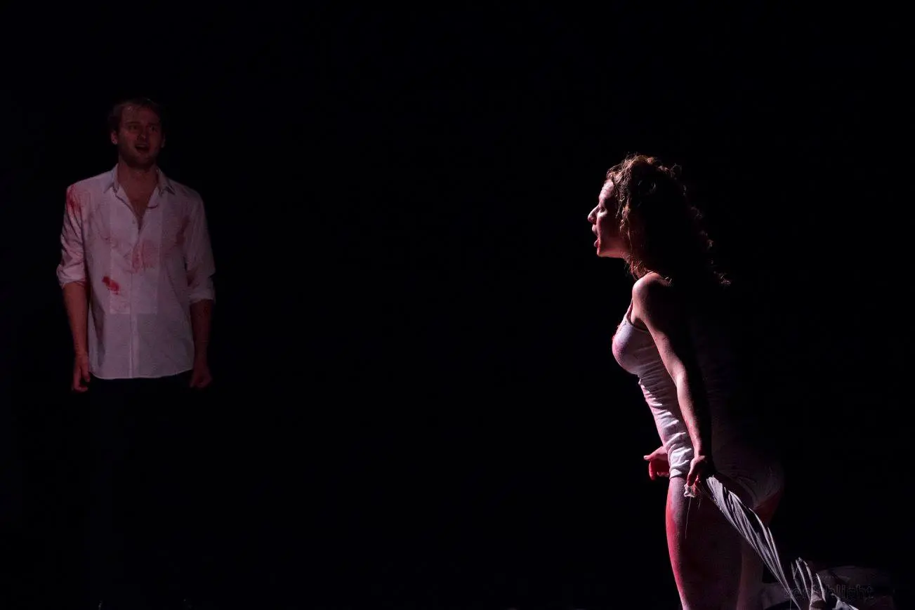 A woman in a white outfit, partially stained with fake blood, stands in profile screaming with intense emotion while holding a piece of cloth. In the dimly lit background, a man in a blood-stained white shirt looks on with a distressed expression.