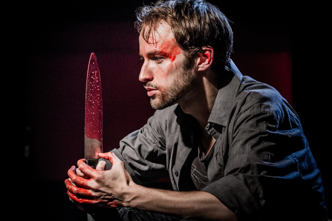 A close-up of an intense actor holding a large blood-stained knife, his face and hands also smeared with fake blood, lit dramatically against a dark background.